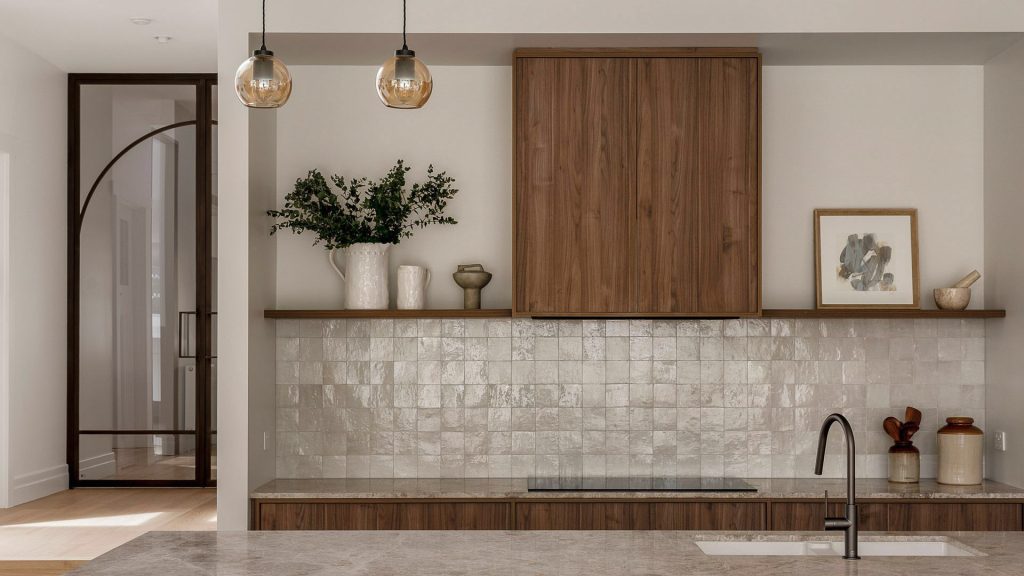 Modern kitchen featuring wooden cabinetry, a textured tile backsplash, and pendant lights, with a glimpse of an arched doorway beyond.