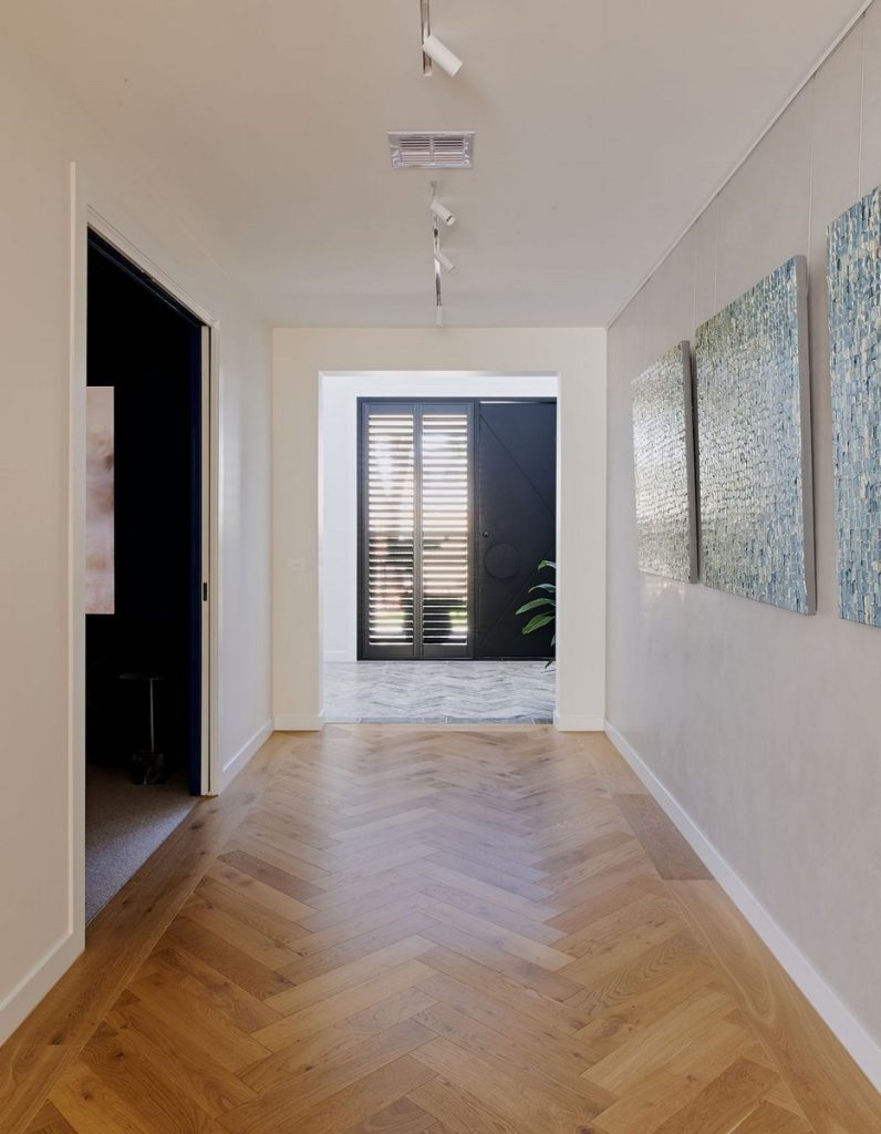 Bright hallway featuring herringbone timber flooring, artwork on the walls, and a view of steel-framed door with louvres and greenery outside.
