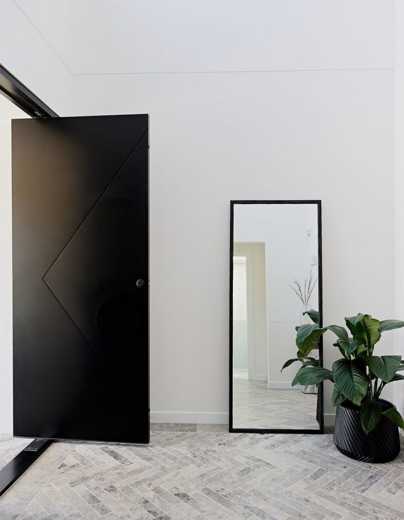 Sleek black door with geometric design, large mirror, and a potted plant against a pale wall, on a herringbone-patterned floor.