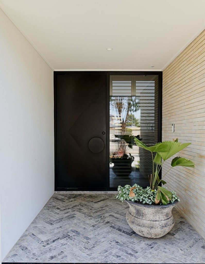 Bespoke black steel door with a circular handle, framed by a light brick wall; potted plant and textured flooring enhance the modern entrance.