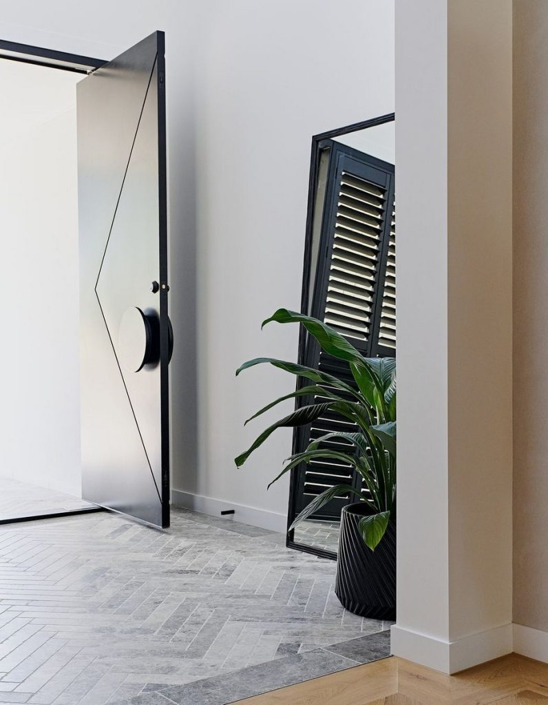 Bespoke black steel door with geometric handle opens to a bright space, featuring a stylish mirror and potted plant on textured flooring.