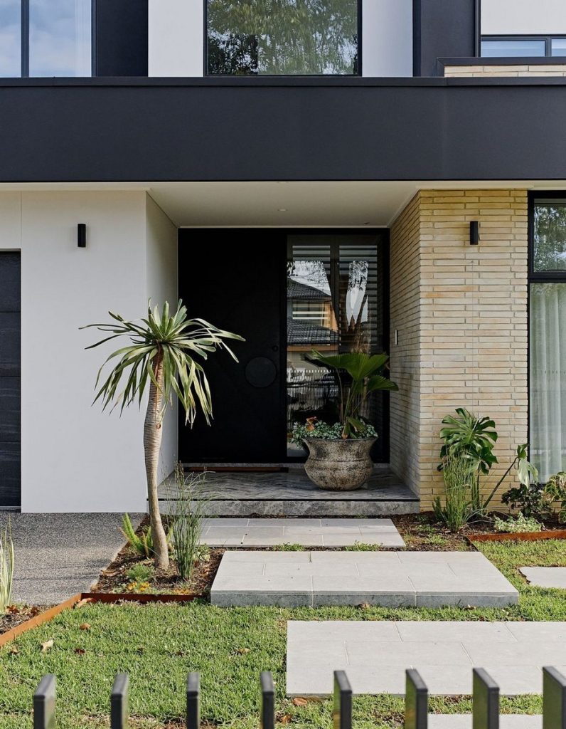 Modern entryway featuring a sleek black door, stone tiles, lush greenery, and a contemporary façade with a mix of materials.