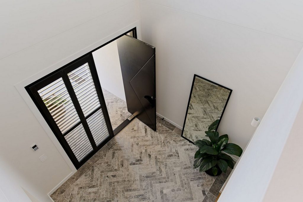Bespoke black steel door open to a light-filled hallway with herringbone patterned flooring, framed mirror, and a large potted plant.