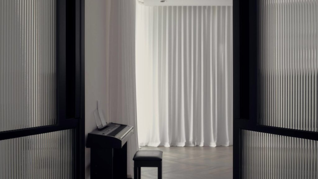 Black steel-framed doors reveal a softly lit interior with sheer white curtains and a piano in the foreground, creating a serene atmosphere.