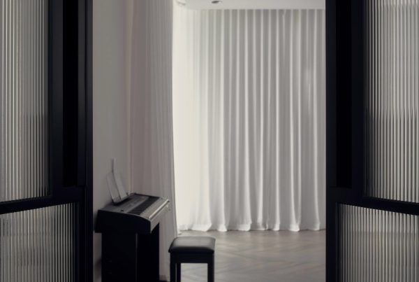 Black steel-framed doors reveal a softly lit interior with sheer white curtains and a piano in the foreground, creating a serene atmosphere.