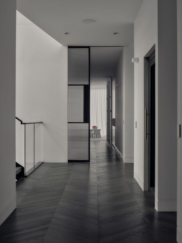 Modern hallway featuring sleek, black-framed glass partitions, light wood flooring, and a minimalist design leading to a softly lit area.