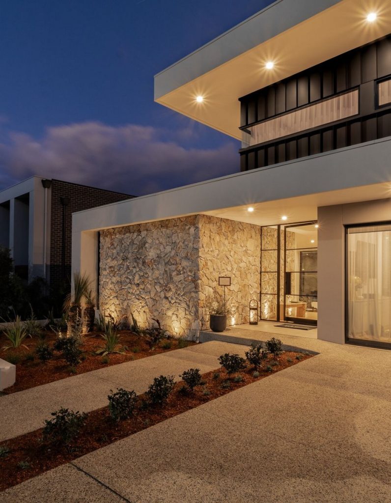 Modern stone house at dusk, featuring a textured stone wall, sleek steel windows, and landscaped pathway illuminated by soft lighting.