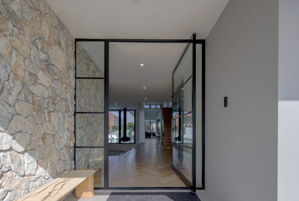 Modern entrance featuring a large glass door framed in black steel, with a stone wall to the left and a wooden bench in the foreground.