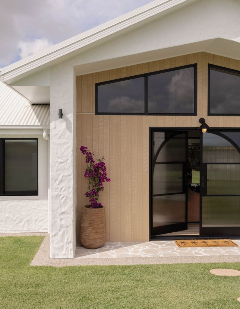 Elegant entrance of Mackay Villa featuring a curved steel-framed door, beige panelling, large black windows, and a potted purple plant.