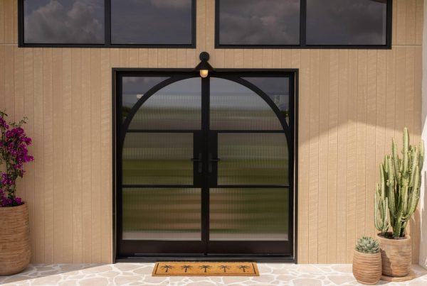 Bespoke steel entry door with an arched design, flanked by potted plants, set against a textured beige wall and large windows.