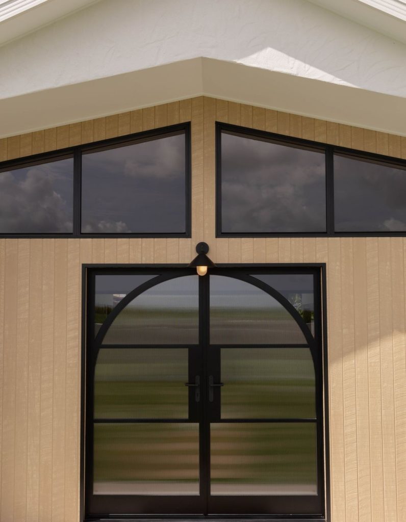 Bespoke steel door with arched top, flanked by large black-framed windows, set against a light-coloured wood-panelled exterior.