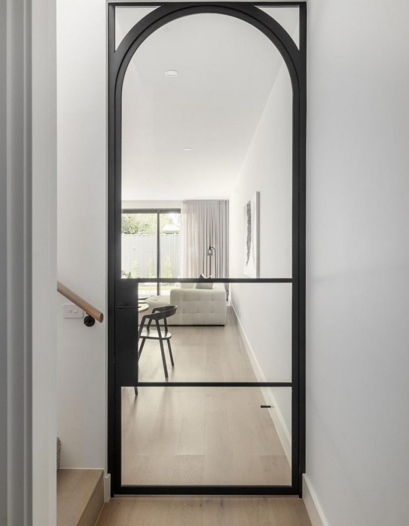 Sleek black steel-framed door with an arched top, leading to a bright living area with a light-coloured sofa and modern decor.