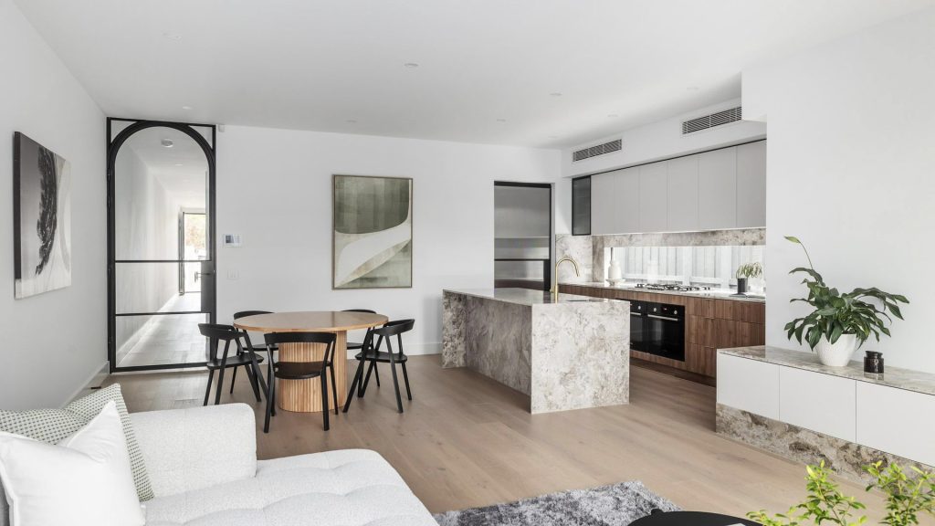 Contemporary open-plan living area featuring a marble kitchen island, dining table, and sleek black-framed glass doors. Soft neutral tones throughout.