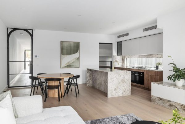 Contemporary open-plan living area featuring a marble kitchen island, dining table, and sleek black-framed glass doors. Soft neutral tones throughout.