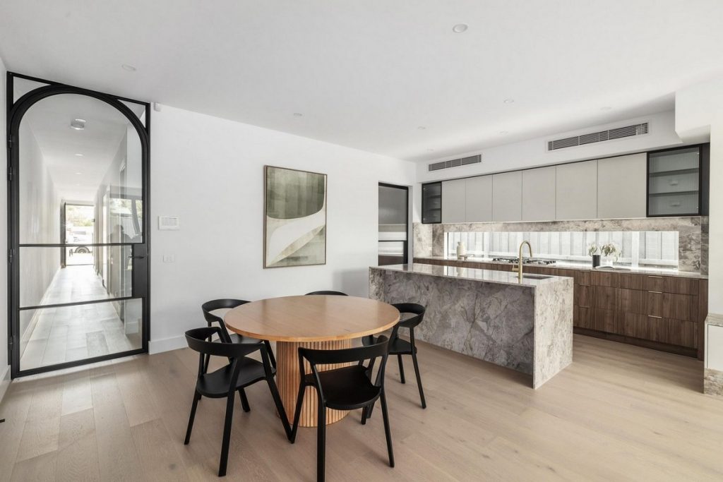 Modern open-plan interior featuring a round wooden dining table with black chairs, sleek kitchen cabinetry, and large windows allowing natural light.