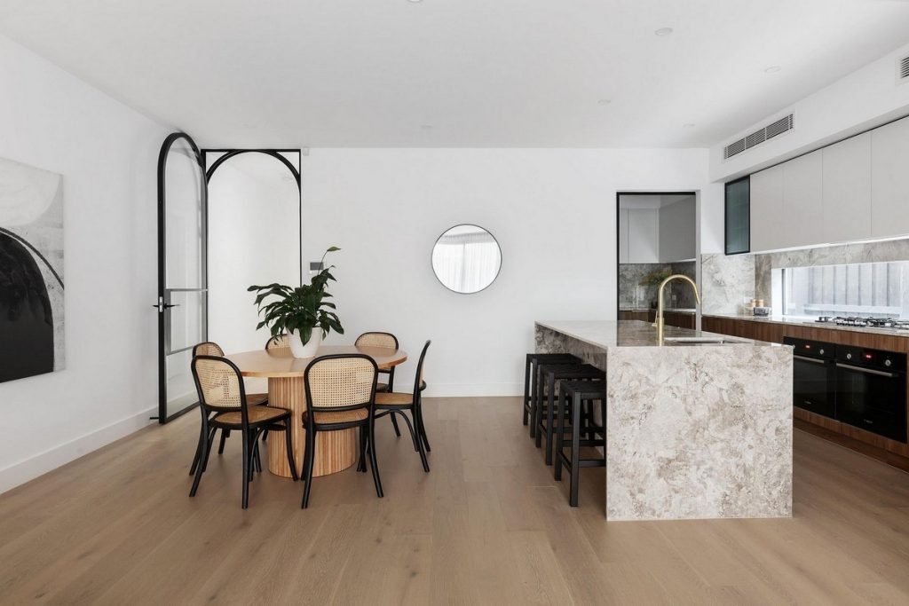Modern open-plan kitchen and dining area featuring a round wooden table, black chairs, stone island, and sleek cabinetry with marble accents.