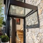 Modern entrance with a sleek black steel door, large glass canopy, and natural stone wall, showcasing inviting interior stairs.