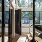Elegant entryway featuring a black steel door, glass walls, and natural light with sleek wooden stairs and greenery visible outside.