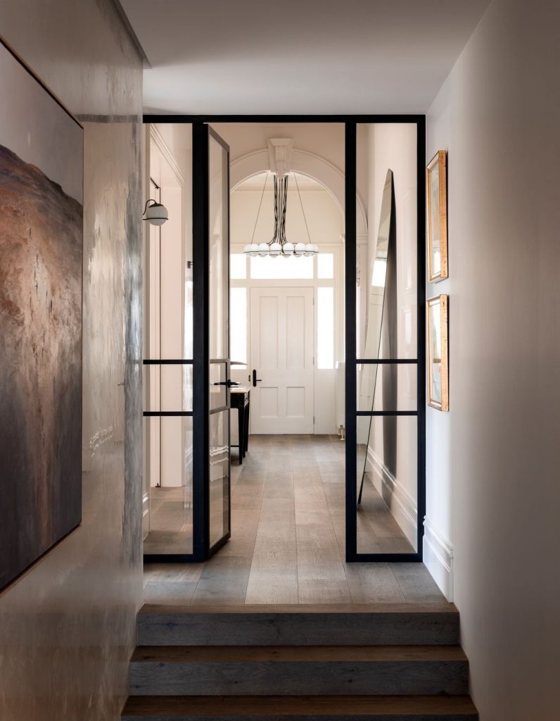 Modern hallway featuring black steel-framed glass doors, natural wood flooring, and elegant wall art, leading to a bright entrance.