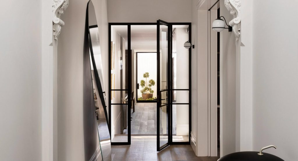 Black steel-framed doors open into a bright hallway, flanked by a large mirror and stylish light fixtures, with greenery visible ahead.