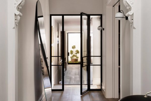 Black steel-framed doors open into a bright hallway, flanked by a large mirror and stylish light fixtures, with greenery visible ahead.