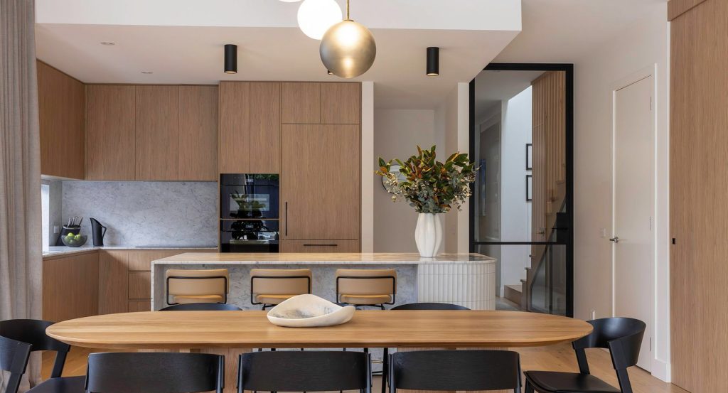 Modern kitchen and dining area featuring wooden cabinetry, a marble island, black chairs, and a striking vase of flowers on the table.