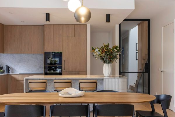 Modern kitchen and dining area featuring wooden cabinetry, a marble island, black chairs, and a striking vase of flowers on the table.