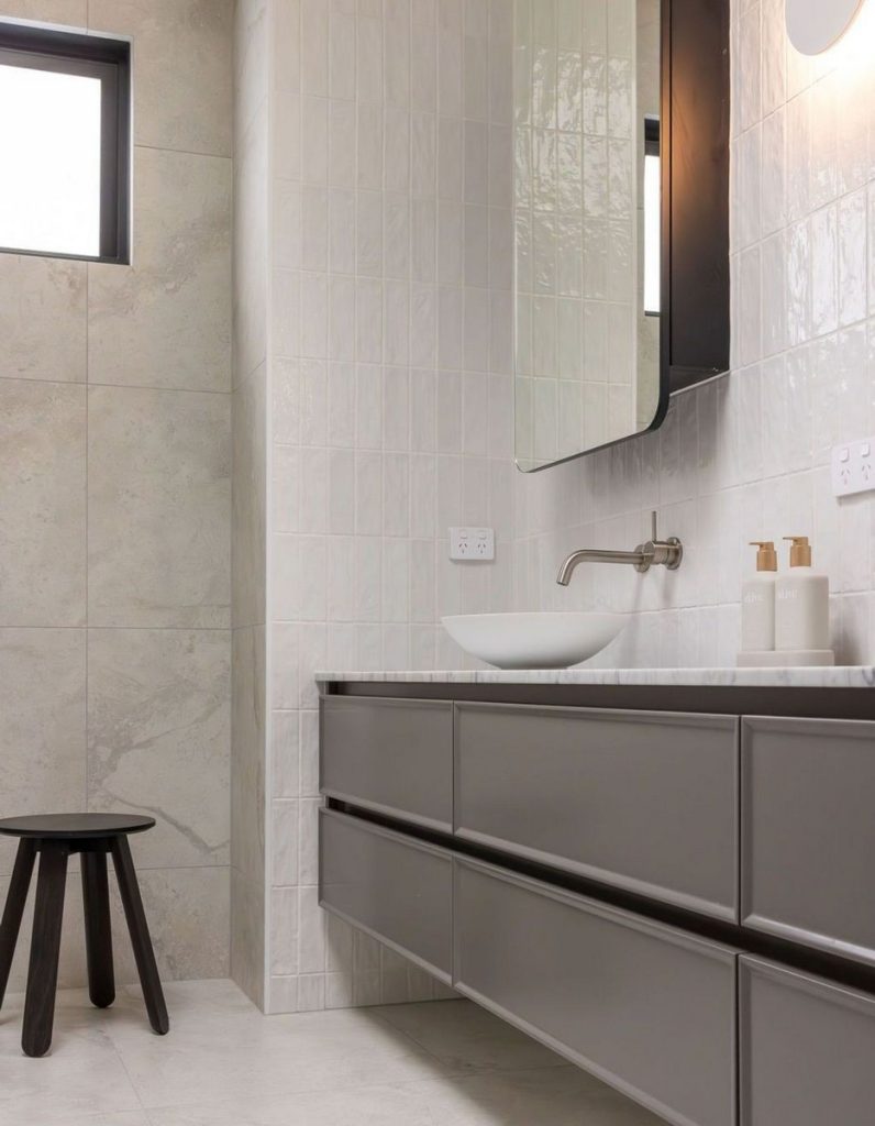 Modern bathroom featuring textured light grey tiles, sleek grey cabinetry, a round basin, and a minimalist mirror, with a wooden stool beside.