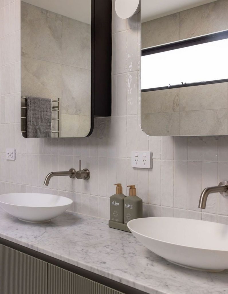 Elegant bathroom with marble countertop, two round basins, modern faucets, and sleek mirrors; neutral tones with grey and white tiles.