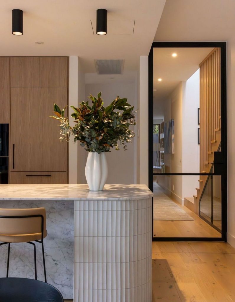 Modern kitchen featuring a curved marble island with a vase of flowers, wooden cabinetry, and a sleek black-framed doorway leading to stairs.