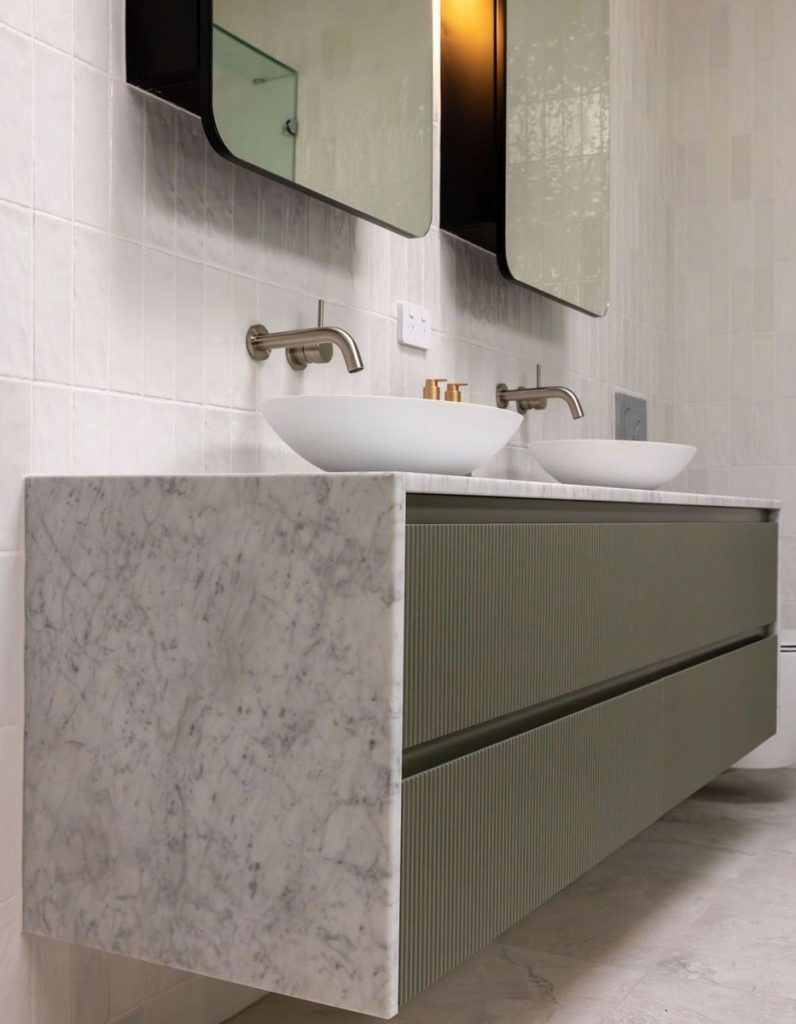 Sleek bathroom vanity with a textured green cabinet, white marble top, and dual basin sinks, set against light tiled walls.