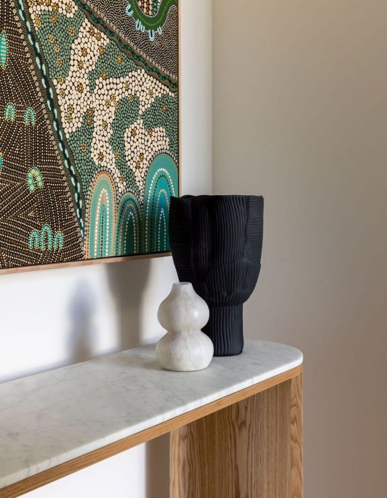 Blackened textured vase and a white sculptural object on a marble-topped wooden console, with a vibrant abstract artwork in the background.