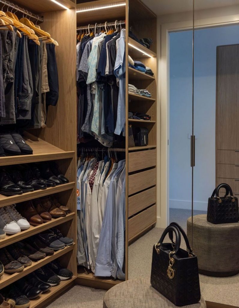 Bespoke walk-in wardrobe featuring wooden shelving, neatly arranged clothes, shoes, and a stylish handbag on a stool.