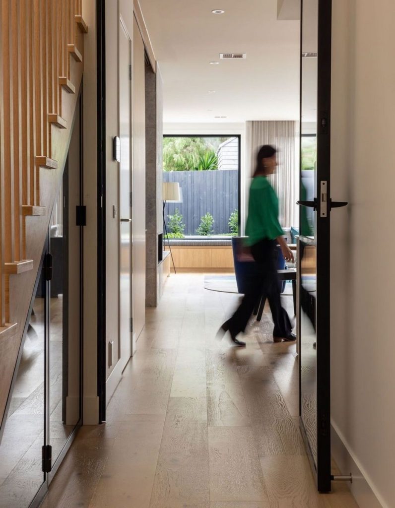 Modern hallway featuring sleek glass doors, wooden stairs, and a blurred figure walking through, with natural light illuminating the space.