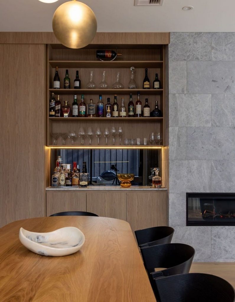 Stylish interior featuring a wooden dining table, a minimalist bar with drinks, and a grey stone wall with ambient lighting.