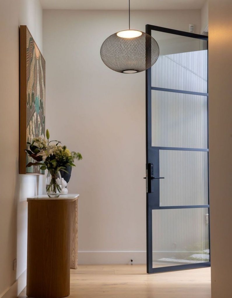 Bright entryway featuring a steel-framed glass door, wood console table, a vase of flowers, and a modern pendant light.