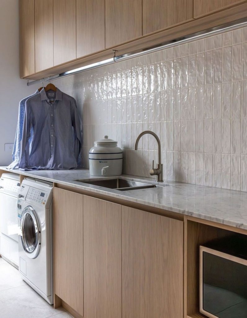Modern laundry space featuring light wood cabinetry, a marble countertop, a stainless steel sink, and a washing machine beside hanging clothes.