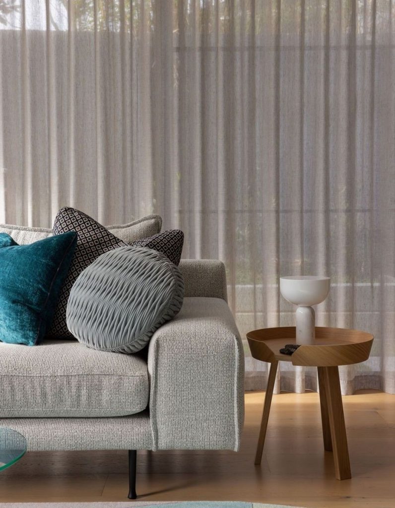 Textured grey sofa adorned with patterned and plush cushions, beside a minimalist wooden side table with a white bowl, against sheer curtains.