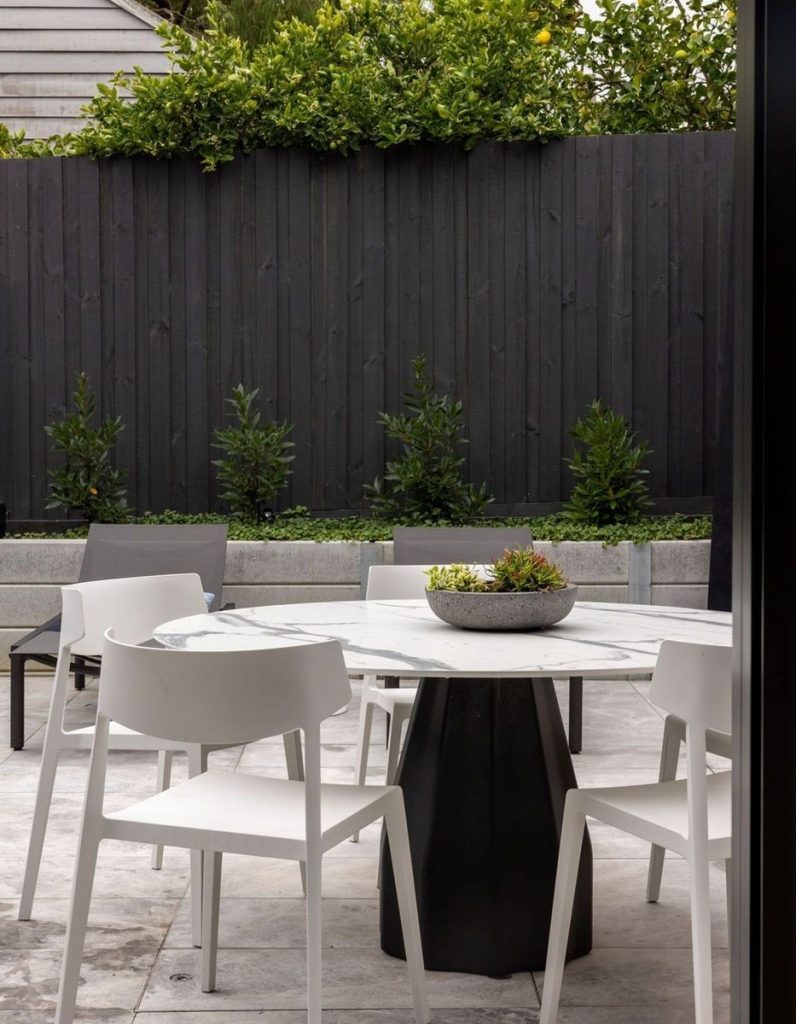 Outdoor dining area featuring a circular marble table with a black base, surrounded by modern white chairs, against a dark wooden fence.
