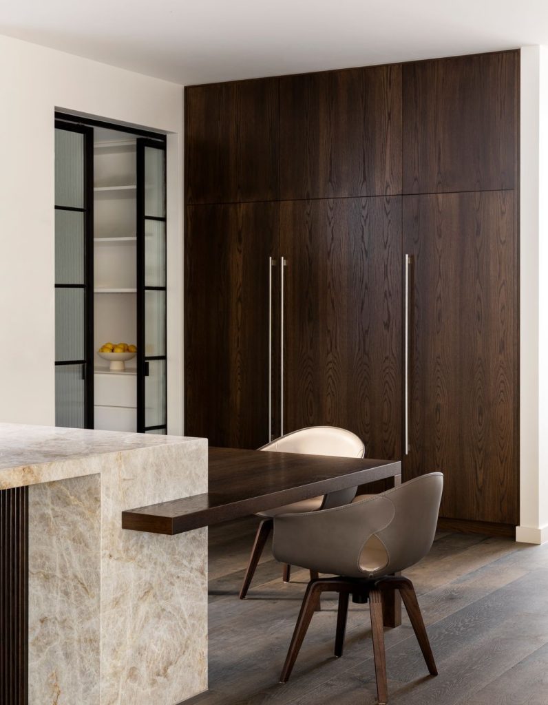 Modern kitchen featuring a sleek wooden cabinet, minimalist steel-framed glass door, and a stylish chair set against warm, neutral tones.