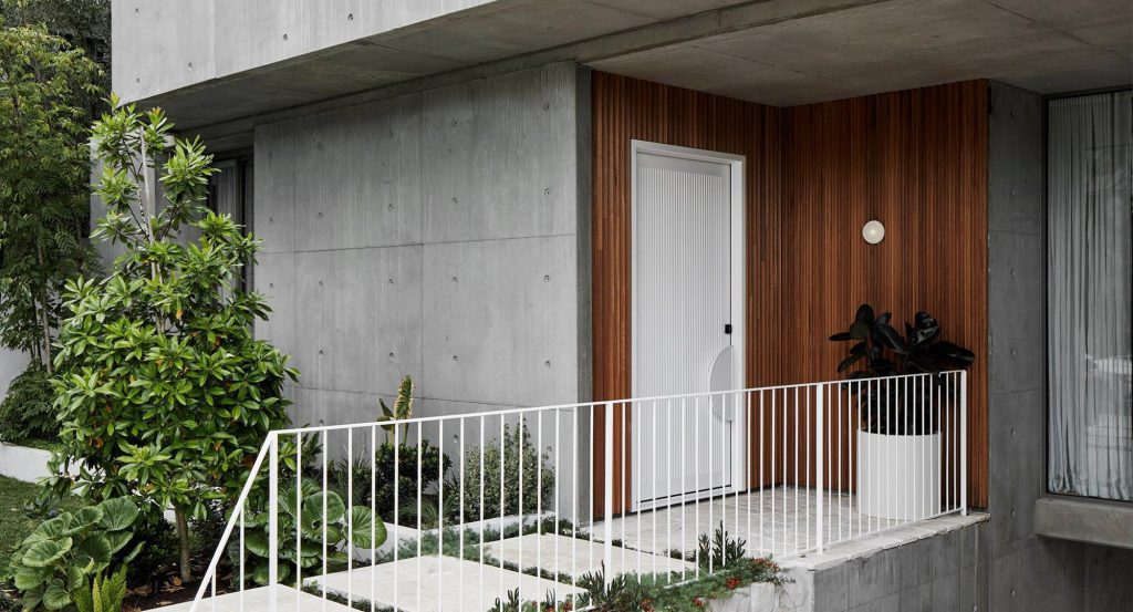 Modern concrete house with a white door, wooden accents, and a planted entrance. Features a white railing and lush greenery.