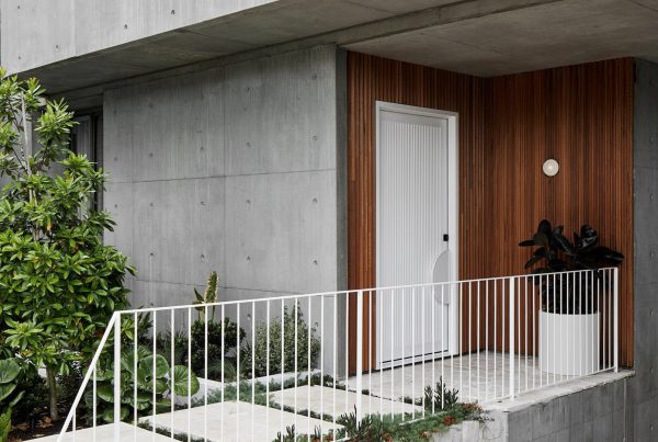 Modern concrete house with a white door, wooden accents, and a planted entrance. Features a white railing and lush greenery.
