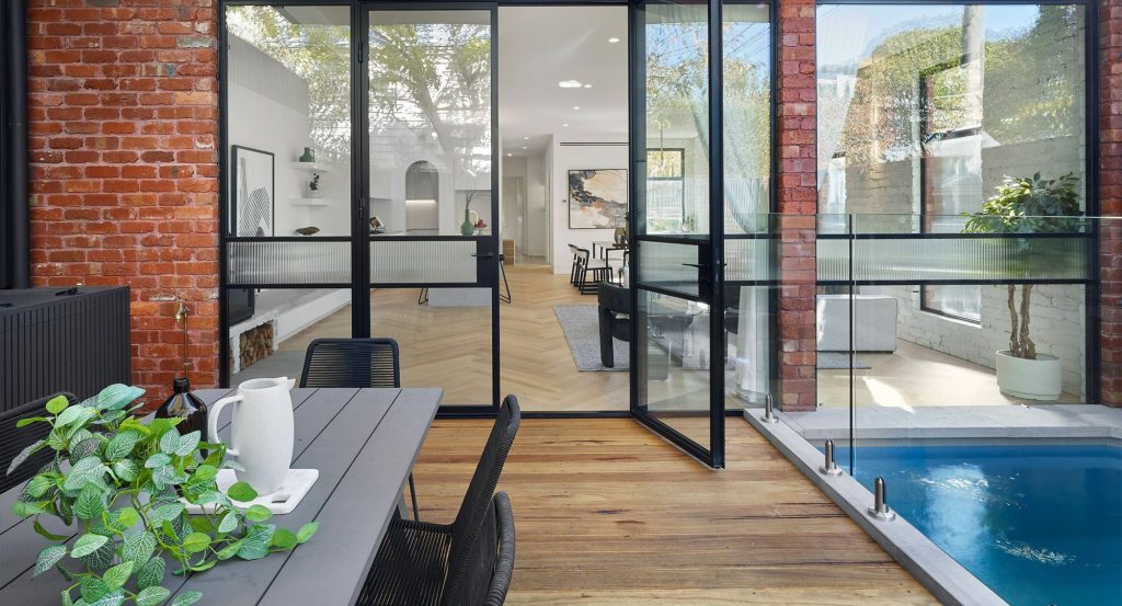 Bespoke steel doors open to a modern interior with a wood and glass layout, featuring a pool and greenery for a seamless indoor-outdoor flow.