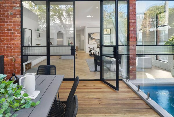 Bespoke steel doors open to a modern interior with a wood and glass layout, featuring a pool and greenery for a seamless indoor-outdoor flow.