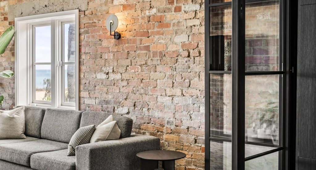 Modern living area featuring a grey sofa, textured brick wall, steel-framed glass door, and a coastal view through a window.