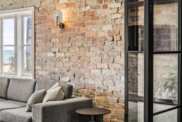 Modern living area featuring a grey sofa, textured brick wall, steel-framed glass door, and a coastal view through a window.