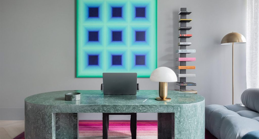 A sleek green marble desk with a laptop, a sculptural lamp, and a colourful book display, complemented by a vibrant abstract artwork.