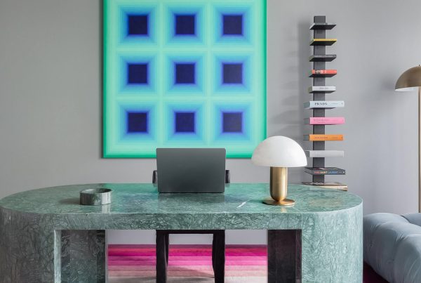 A sleek green marble desk with a laptop, a sculptural lamp, and a colourful book display, complemented by a vibrant abstract artwork.