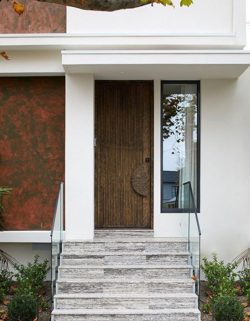 Textured bronze door with circular handle, framed by large glass panel. Steps lead up to a modern facade with earthy tones.