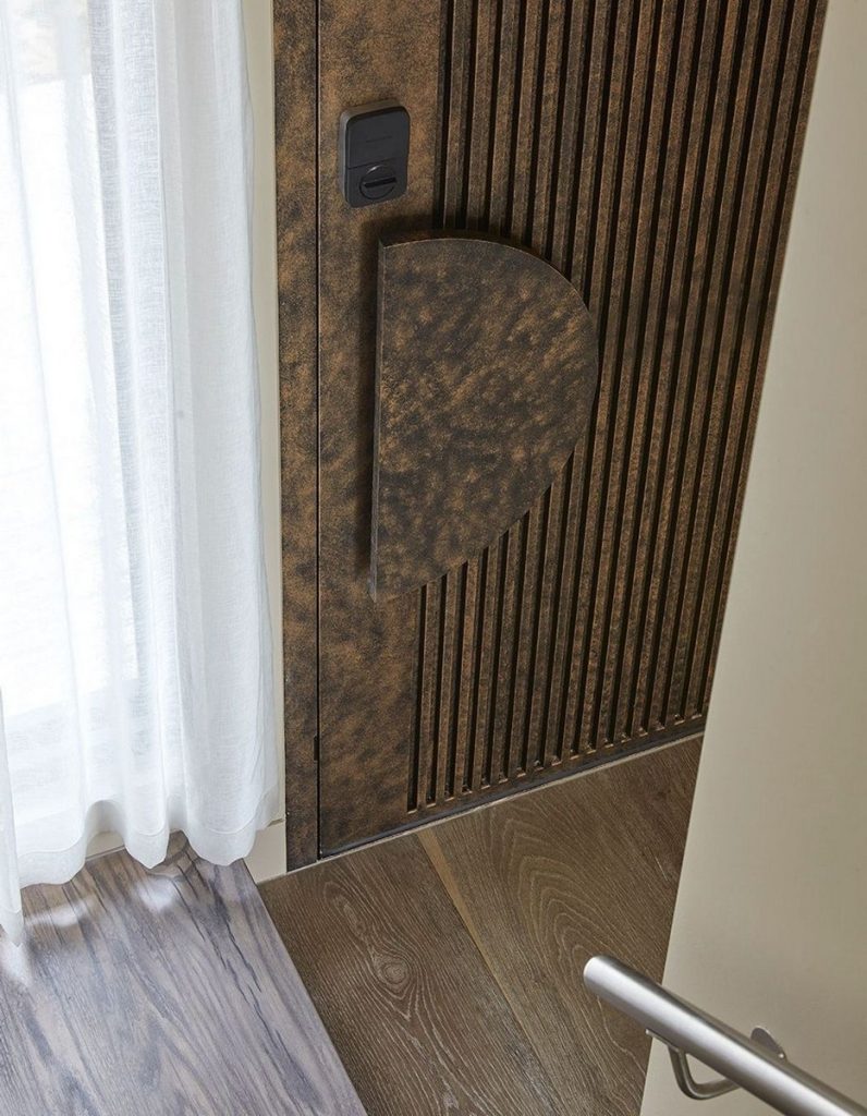 Textured bronze door with a distinctive curved handle, complemented by wooden flooring and sheer white curtains, creating a modern interior.
